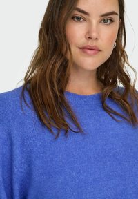 Donna con capelli marroni ondulati, indossa piccoli orecchini ad anello e un maglione blu lavorato a maglia, su uno sfondo bianco semplice.