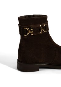 Braune Wildlederschuh mit einem goldfarbenen D-förmigen Schnallen- und Riemen-Detail. Flacher Absatz mit glatter Oberfläche, strukturierter Obermaterial und runder Zehenpartie.