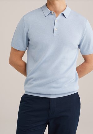 Mand iført en lyseblå kortærmet polo shirt og mørkeblå bukser, stående med hænderne bag ryggen mod en neutral baggrund.