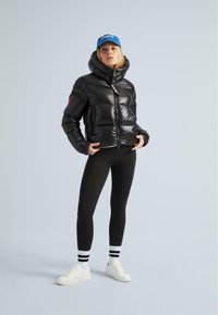 Donna che indossa un giubbino imbottito nero lucido, leggings neri, sneakers bianche, calze a righe e un cappellino blu su uno sfondo semplice.