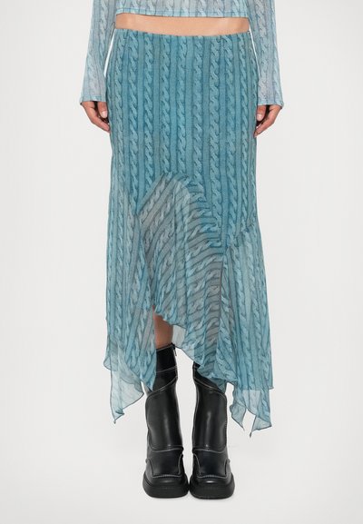 Mujer viste una falda azul transparente de punto trenzado con dobladillo asimétrico sobre pantalones cortos a juego, combinada con botas de cuero negras hasta medio muslo.