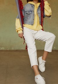Chaqueta de mezclilla con diseño corto, sudadera amarilla, pantalones blancos y zapatillas blancas. Flecos rojos que acentúan el atuendo.