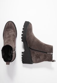 Hnědé semišové kotníkové boty s masivní černou gumovou podrážkou, bočním zipem a textilním poutkem na zadní straně pro snadné obouvání.