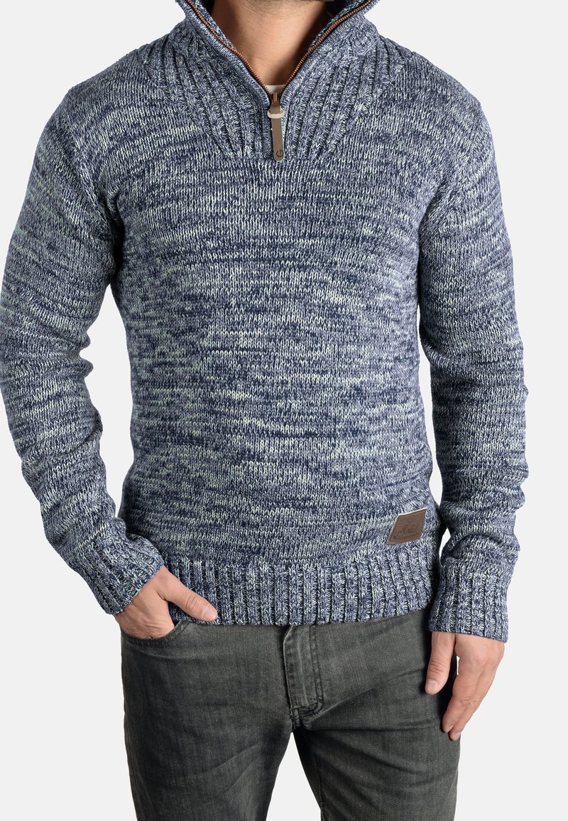 Homme portant un pull tricoté à motifs bleus et blancs avec une fermeture éclair partielle et des poignets côtelés, associé à un pantalon gris foncé, les mains dans les poches.