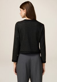 Chaqueta de tweed negra con mangas largas, tela texturizada y un diseño crop. Acentuada con un dobladillo en forma de ondas y combinada con pantalones grises.
