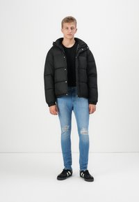 Schwarze Pufferjacke mit Kapuze, die ein gestepptes Design und Druckknöpfe aufweist. Getragen über einem schwarzen Shirt und kombiniert mit hellblauen, zerrissenen Jeans und schwarzen Schuhen.