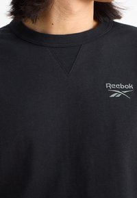 Svart bomullströja med V-hals och en diskret Reebok-logotyp i mörkgrått på vänster bröst. Slät textur.