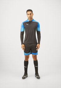 Schwarzes Sporthemd mit blauen Farbverlaufärmeln, kombiniert mit schwarzen Shorts mit blauen Akzenten. Schwarze Kniestrümpfe und Fußballschuhe.