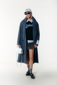 Long grey pinstriped coat, black sweatshirt with "Rebel" text, sequined shorts, white handbag, grey socks, black loafers, and blue cap.