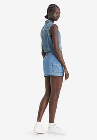 Gilet in denim a pantaloni cropped con colletto, abbinato a pantaloni shorts in denim a vita alta. Colore azzurro chiaro, con tasche e sneakers bianche.