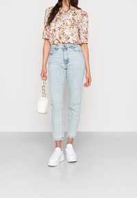 Blus med blommönster och puffärmar, högmidjade ljus denimjeans och vita plattformssneakers. Håller i en liten vit väska.