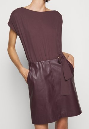 Femme portant une robe couleur prune avec une ceinture en tissu nouée et une jupe en simili cuir, les mains dans les poches, sur un fond blanc uni.