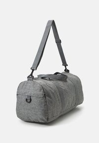 Bolsa de viaje de tela gris con forma redondeada, que cuenta con asas en la parte superior y una correa de hombro ajustable. Incluye un bolsillo lateral con hardware de bucle.