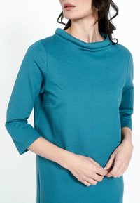 Kvinna i en teal-färgad klänning med trekvartsärmar och upplagd krage, med händerna knäppta vid midjan och tittar åt sidan mot vit bakgrund.