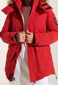 Rode parka met een bontgevoerde capuchon, ritssluiting, klittenbandaccenten en een voorzak. Kenmerkt zich door een gestructureerde stof en contrasterende mouwen.