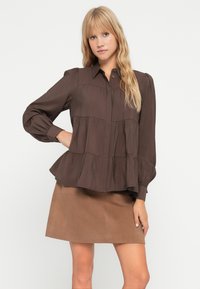Bruine blouse met lange mouwen, knoopsluiting aan de voorkant en een gelaagd ontwerp. De stof is soepel en heeft een relaxte pasvorm, gecombineerd met een tan kleurige rok.