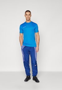 Blaues Sportshirt mit kurzen Ärmeln, kombiniert mit blauen Jogginghosen mit lila Akzenten und orangefarbener Paspelierung. Schwarze Schuhe runden das Outfit ab.