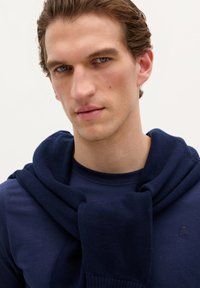 Ung mand med blå øjne iført en marineblå skjorte og en marineblå sweater draperet over skuldrene mod en ensfarvet hvid baggrund.