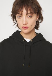 Sudadera con capucha negra, con un bolsillo delantero tipo pouch, acentos de joyería en tono dorado, cordones y tejido suave y texturizado.