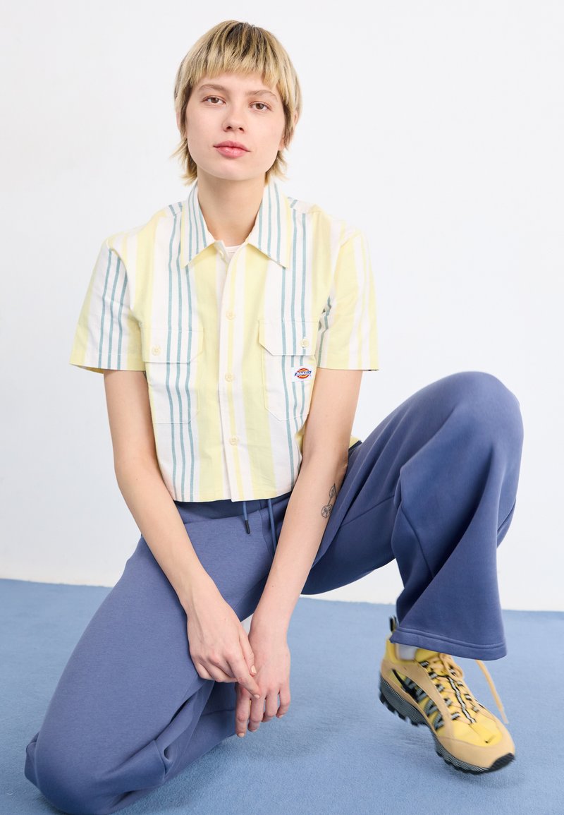 Kurzärmliges Hemd mit Knopfleiste in pastellgelben und blauen Streifen, kombiniert mit lockeren blauen Sweatpants und gelben Sneakers mit schwarzen Akzenten.