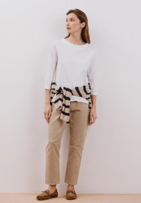 Top bianco a maniche lunghe con dettagli di bottoni, abbinato a pantaloni beige e una cintura a righe in maglia, completato da scarpe mocassino marroni.