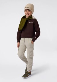 Bruine trui met logo, beige muts, groene sjaal, lichtgrijze cargobroek en olijfgroene sneakers met witte zolen.