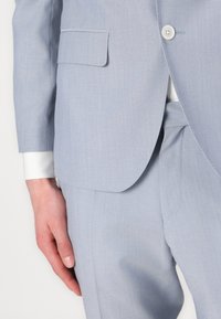 Traje azul claro hecho de una tela texturizada, con cierre de un solo botón y un bolsillo en el pecho. Puño de camisa blanca lisa visible.