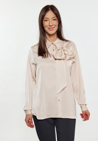 Blusa lunghezza maniche in beige chiaro in tessuto setoso, caratterizzata da una chiusura con bottoni frontale, colletto e un grande dettaglio a fiocco sullo scollo.
