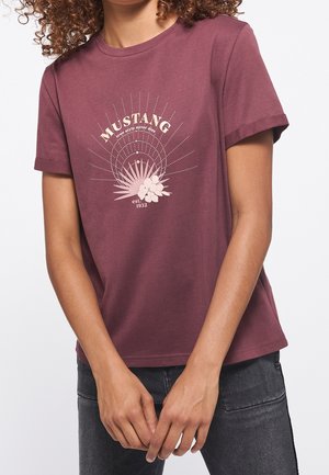 Femme portant un t-shirt violet avec des manches retroussées et un graphique "Mustang" avec un motif de soleil et de fleurs imprimé sur la poitrine.