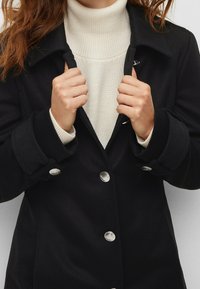 Donna che indossa un cappotto nero con bottoni argentati sopra un maglione a collo alto bianco in knit, mentre tiene il colletto del cappotto con entrambe le mani.