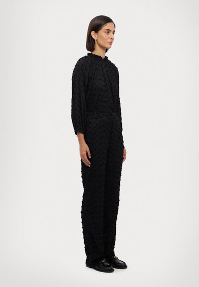 Black jumpsuit with textured fabric, long sleeves, and a high collar. Features a belted waist and wide-leg design, paired with black shoes.