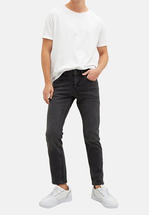 Person iført hvid kortærmet skjorte, sorte slim-fit jeans og hvide sneakers, stående med den ene hånd i lommen mod en hvid baggrund.