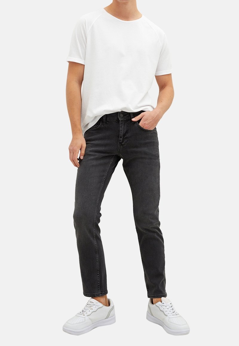 Personne portant une chemise blanche à manches courtes, un jean noir slim et des baskets blanches, debout avec une main dans la poche, sur un fond blanc.