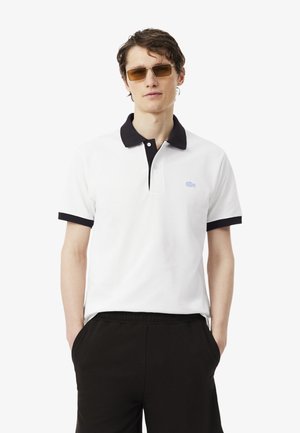 Homme portant un polo blanc à manches courtes avec col noir et bords de manches noirs, pantalon noir et lunettes de soleil rectangulaires marron.
