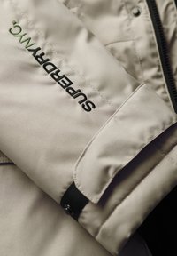 Manche de veste beige avec logo "SUPERDRYMC" brodé en noir et vert et une sangle à bouton-pression noire près du poignet.