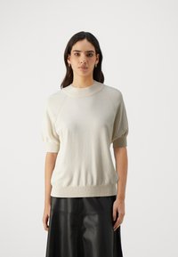 Lys beige strikkegenser med rund hals, korte puffermarmer og ribbestrikket kant. Kombinert med et svart skinnskjørt for kontrast.