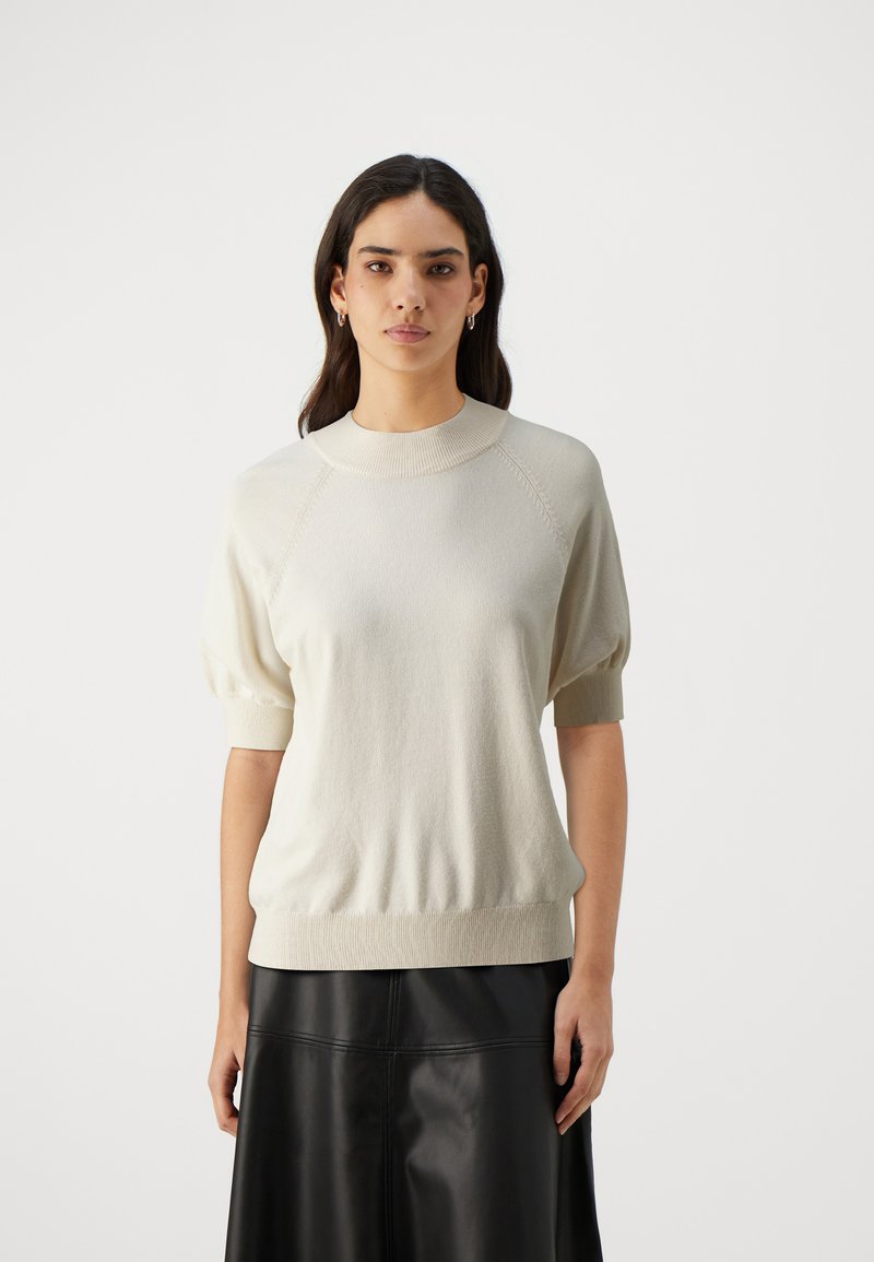 Lys beige strikkegenser med rund hals, korte puffermarmer og ribbestrikket kant. Kombinert med et svart skinnskjørt for kontrast.