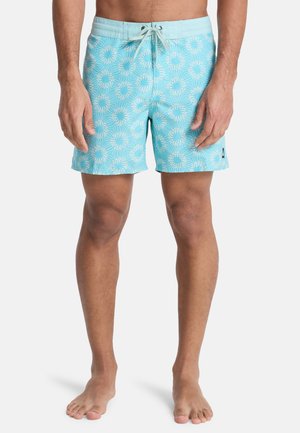 Mężczyzna w jasnoniebieskich kąpielówkach z białymi wzorami promieni słonecznych, związaną talią, stojący boso na jednolitym tle.