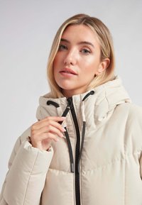 Chaqueta de plumas beige con cuello alto, cremallera negra y cordón. Presenta una textura acolchada y detalles cosidos.