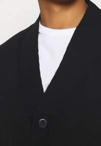 Svart kardigan med V-ringad design, med ribbat mönster och en enda rund svart knäppning, bärs över en vit t-shirt.