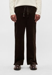 Brūnas kordurojas bikses ar brīvu piegriezumu, kas aprīkotas ar aukliņu jostu un redzamu riboto tekstūru. Kombinētas ar tumšiem apaviem.