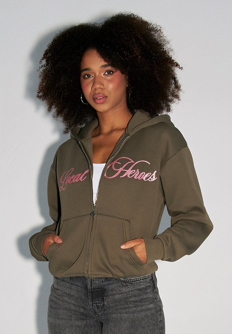Oliwkowa bluza z kapturem na zamek z napisem "Local Heroes" w różowym kursywie. Posiada kaptur i kieszenie z przodu. Gładki materiał i luźny krój.