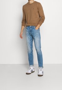 Brauner Langarmpullover, hellblaue Jeans, weiße Schuhe mit schwarzen Streifen. Lässige Freizeitkleidung mit lockerer Passform und minimalistischem Design.