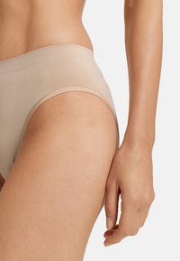 Beige saumatonta alusvaatetta korkealla vyötäröllä, pehmeällä pinnalla ja sileällä viimeistelyllä, korostaen minimalistista muotoilua. Käsi lepää vierellä.