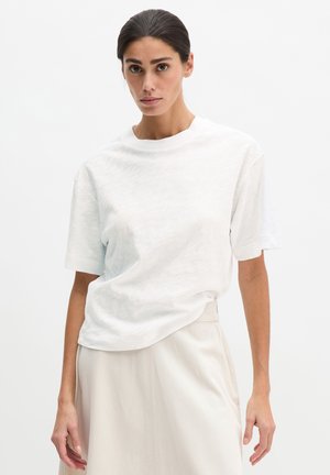 Woman with dark hair in a white short-sleeve T-shirt and beige skirt, standing against a plain white background.