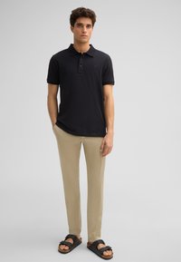 Schwarzes Poloshirt mit einer Knopfleiste mit drei Knöpfen, beigen Hosen und schwarzen Sandalen mit einem Riemen mit Schnalle. Glattes Material und lässige Passform.