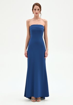 Donna che indossa un abito blu senza spalline, lungo fino a terra, e sandali aperti, in piedi davanti a uno sfondo chiaro e semplice.