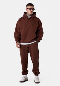 Hoodie marron avec une poche frontale, coupe décontractée et joggers à cordon. Le tissu semble doux ; présente un branding minimal. T-shirt blanc en dessous.