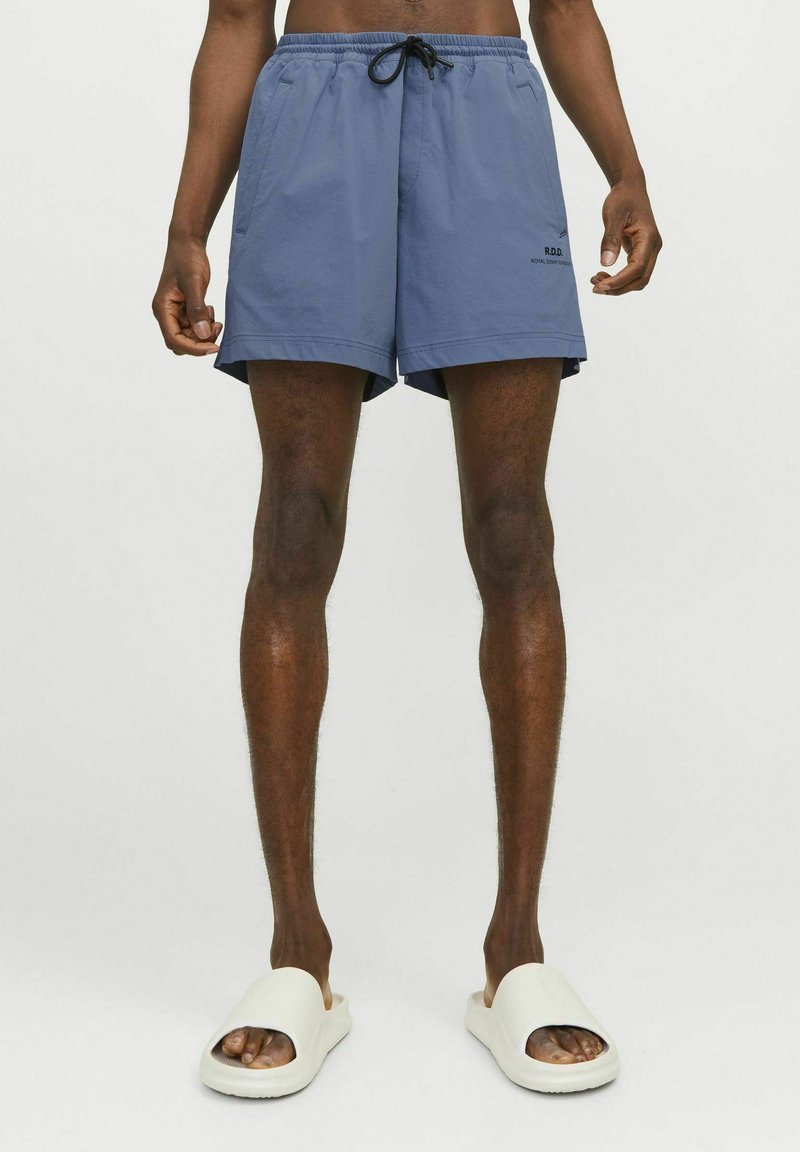 Shorts de bain bleus avec une taille élastique, cordon de serrage, poches latérales et logo R.D.C. sur le devant. Portés avec des sandales blanches.