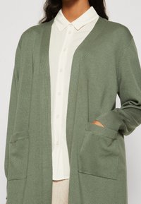 Kvinna iklädd en ljusgrön öppen cardigan med fickor över en vit skjorta med knappar och krage.